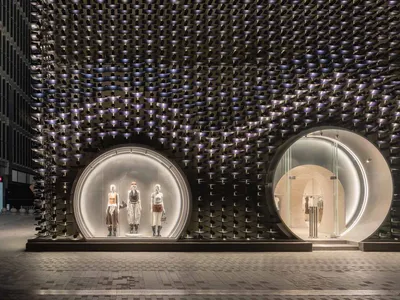 Exterior night view of a luxury retail building featuring a dramatic black perforated metal facade with two large illuminated circular entrances displaying mannequins.