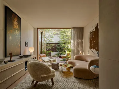 Modern living room featuring a curved boucle sofa, textured boucle armchair, stone coffee table, large abstract paintings, and an open view to a lush green courtyard garden.