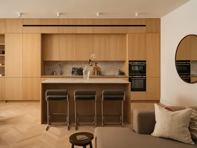 Modern open-plan kitchen featuring light oak cabinetry, a marble island with three dark green bar stools, herringbone wood flooring, and a neutral sofa in the foreground.