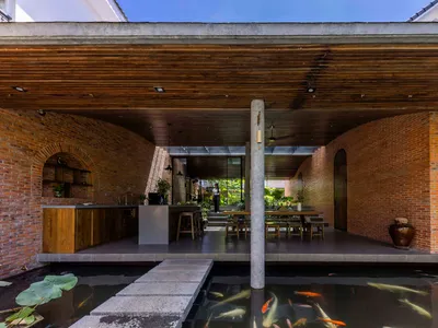 Modern tropical courtyard house featuring curved exposed brick walls, wooden ceiling, open kitchen, dining area, and a foreground koi pond with stepping stones.