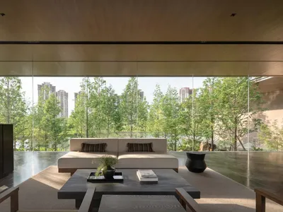 現代簡約客廳，設有低矮米色沙發、深色石材茶几、木質天花板，以及可眺望茂密綠樹與城市建築的落地玻璃窗。