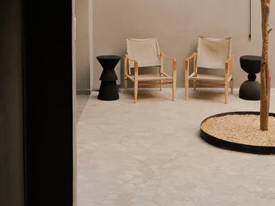 View through a dark arched doorway into a minimalist concrete courtyard featuring two wooden canvas sling chairs, black sculptural side tables, and a young tree planted in a circular gravel bed.
