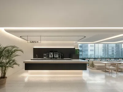 Modern minimalist cafe interior featuring a long dark wood service counter with white stone top, illuminated from below, a large potted palm tree in the foreground, and light wood seating area in the background.
