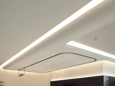 Modern minimalist cafe interior featuring a long dark wood service counter with white stone top, underlit with warm LED lighting, white tiled floor, and dramatic linear ceiling lights.