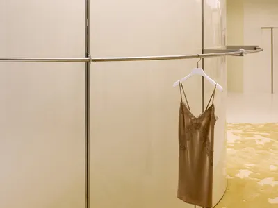 Curved high-gloss cream lacquered wall in a boutique with a brown silk slip dress hanging on a white hanger, set on a textured chartreuse carpet.