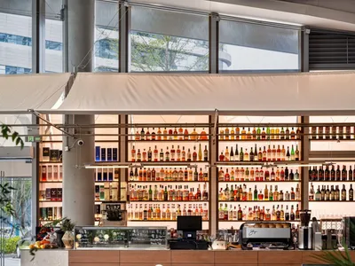 Modern cafe interior with round wooden tables, brown velvet chairs, and a striking illuminated wall of liquor bottles behind a walnut bar counter.