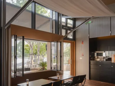 Modern cafe interior featuring a large retractable beige fabric awning, corten steel details, wooden tables, black folding chairs, and full-height glass doors opening to an outdoor terrace.