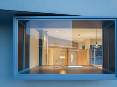 透過大型藍色鋼框窗戶看到的極簡精品店內部，包含橡木櫃體、混凝土牆面，以及擺放現代檯燈的深色展示桌。