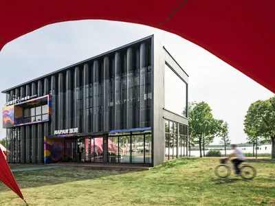 現代兩層商業建築，外牆採用深色穿孔金屬，搭配捷安特與那帕的彩色圖形品牌，坐落於湖畔，前景有單車騎士經過。