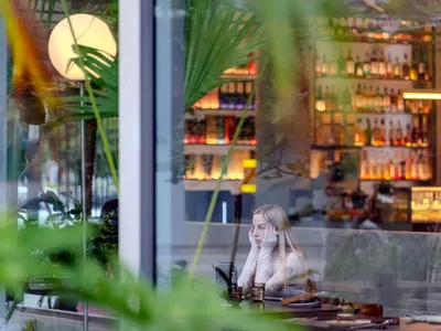 A young woman with long blonde hair sitting at a modern restaurant bar counter, viewed through large glass windows and tropical plants, with illuminated backlit bottle shelves in the background.