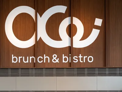 Modern brunch bistro interior featuring a large walnut wood wall with white logo, concrete lower walls, built-in shelving, and round dining tables with mauve chairs.