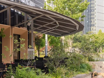 Modern restaurant outdoor seating area featuring a large curved corten steel canopy, black furniture, and abundant greenery with modern buildings in the background.