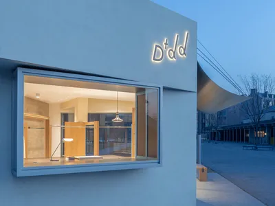 Exterior view of a modern minimalist concrete building with a large illuminated display window showing warm wooden interior elements and a hanging pendant lamp at dusk.