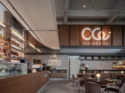 Modern brunch bistro interior with walnut wood counter, brown velvet chairs, round dining tables, and large illuminated logo on the back wall.