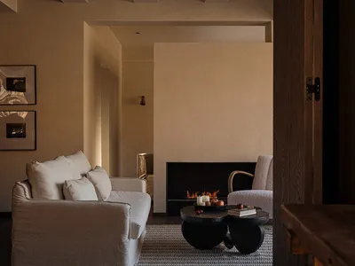 Modern minimalist living room featuring a white linen sofa, sculptural black coffee table, lit fireplace, and textured beige plaster walls with exposed wooden ceiling beams.