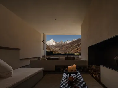 Minimalist bedroom interior with built-in daybed, modern fireplace, textured plaster walls, and large window overlooking snow-capped mountain range.