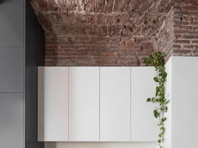 Modern home office nook featuring a white built-in desk, black leather office chair, exposed brick ceiling, and trailing green plants.