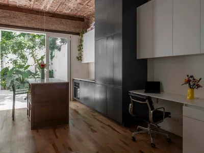 Industrial loft interior featuring exposed brick ceiling, dark grey kitchen cabinetry, white office desk with black leather chair, and large glass doors opening to a sunny garden.