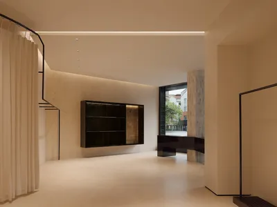 Modern minimalist living room featuring travertine stone walls, a floating dark wood shelving unit with integrated lighting, sheer curtains on a curved black rail, and a view to an outdoor courtyard.