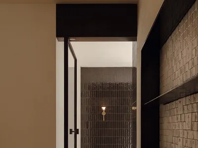 View from a neutral hallway through a black-framed glass door into a shower room lined with dark textured tiles and a brass wall sconce.
