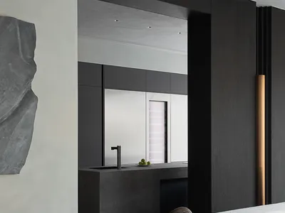 Modern minimalist living room featuring dark concrete walls, a large abstract stone sculpture on the left, sculptural bronze artwork on a black pedestal, and a view into a concrete kitchen with a sleek faucet.