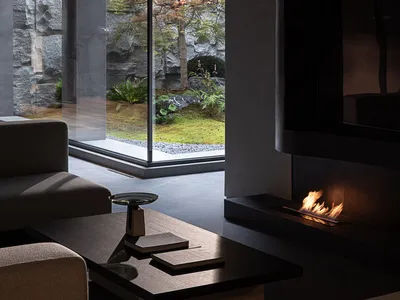 Modern minimalist living room interior featuring raw concrete walls, a linear fireplace, low-profile neutral sofas, and a view of a Japanese-style garden with maple tree and stone wall.