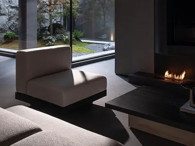 Modern minimalist living room interior featuring raw concrete walls, a built-in linear fireplace, low-profile beige fabric sofa, and a tranquil Japanese garden visible through floor-to-ceiling glass windows.