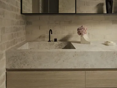 Modern bathroom featuring a solid travertine floating vanity with integrated sink, matte black faucet, light wood cabinetry, and textured beige brick walls with a large frameless mirror.