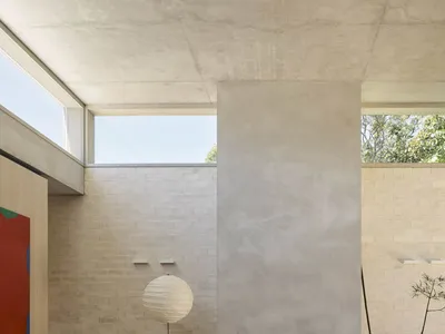 Modern minimalist living room featuring a large concrete fireplace wall, oak lattice coffee table on a cream rug, wooden bench, paper floor lamp, and natural daylight from high windows.
