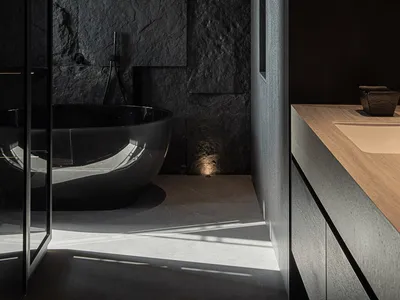 Modern dark minimalist bathroom featuring a glossy black freestanding tub against a textured natural stone wall, paired with a light wood floating vanity and dramatic wall sconce lighting.