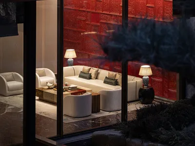 Night view through glass doors into a luxurious contemporary Chinese living room with a dramatic red lacquered carved wall, cream sofas, and elegant lighting.
