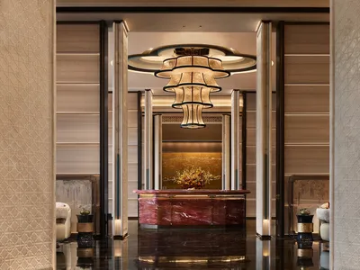 Elegant hotel lobby featuring a striking red marble reception desk, a large multi-tiered chandelier, glossy dark floor, and beige patterned wall panels.