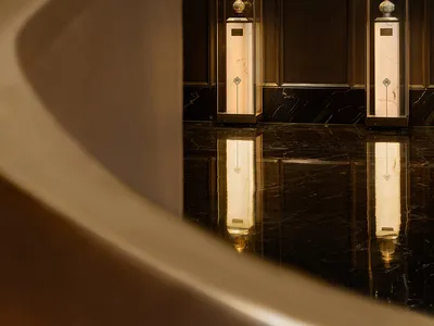 A sophisticated luxury lobby featuring a sweeping curved marble wall with integrated lighting, paired with two illuminated glass display cases showcasing ceramic vases on marble pedestals, all reflected in a highly polished dark marble floor.