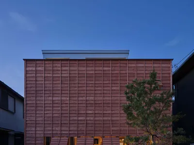 現代兩層杉木住宅，水平木板外牆搭配溫暖燈光的折疊木遮陽板，黃昏時分，周圍環繞著極簡風格的礫石庭園與一棵小松樹。