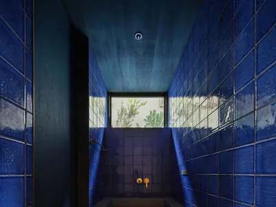 Narrow utility bathroom entirely clad in glossy cobalt blue tiles with a raw concrete sink and cabinet under a high window showing green foliage outside.