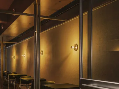Narrow restaurant dining area with black tables and chairs lined against a textured wall, illuminated by warm brass wall sconces creating dramatic lighting and long shadows on a dark tiled floor.