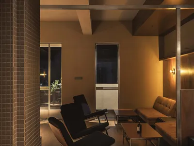Modern brutalist-style lounge featuring two black lounge chairs with wooden frames, a brown fabric sofa, concrete structural pillars, warm wood paneling, and layered ambient lighting at night.