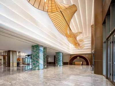 Modern luxury hotel lobby interior with polished marble floors, green marble pillars, and a massive undulating wooden chandelier hanging from a white coffered ceiling.