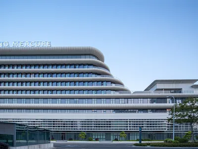 Exterior view of the Grand Mercure hotel featuring a modern curved, multi-layered white facade with extensive glass windows under a clear blue sky.