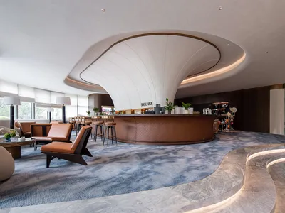 Modern luxury hotel lounge featuring a large curved quilted brown leather bar, sculptural white ceiling canopy, blue patterned carpet, leather armchairs, and illuminated marble steps.