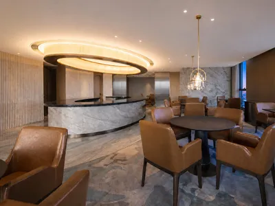 Modern luxury hotel lounge featuring a curved marble bar, cognac brown leather armchairs, round dining tables, and a striking circular pendant light fixture with warm ambient lighting.