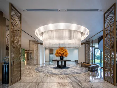 Spacious luxury hotel lobby with polished marble floors, a massive circular tiered chandelier, golden decorative room dividers, and a central round table with orange floral arrangement.