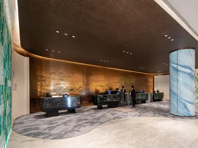 Modern luxury hotel lobby with dark green marble reception desks, a large cylindrical blue marble pillar, golden Chinese landscape metal wall art, and green glass mosaic accent walls.