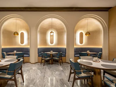 Modern restaurant dining area with three illuminated arched alcoves containing blue banquette seating, round marble tables, and teal upholstered wooden chairs under pendant lights.
