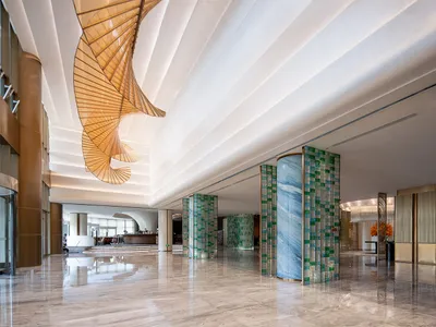 Spacious luxury hotel lobby with polished marble floors, blue and green mosaic pillars, and a large golden spiral chandelier hanging from a coffered ceiling.