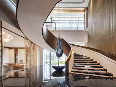 Double-height luxury lobby featuring a dramatic curved black marble staircase, a large teardrop pendant light, and reflective polished marble floors.