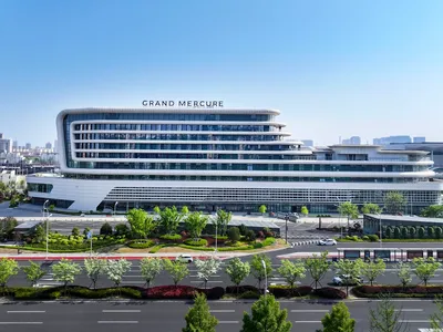 Front view of the modern Grand Mercure hotel building with distinctive curved white facade, extensive glass windows, and landscaped garden under a clear blue sky.