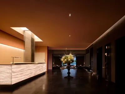 Modern luxury hotel lobby featuring a long illuminated onyx reception desk, dark stone flooring, a central round table with large floral arrangement, and warm terracotta ceiling with recessed lighting.