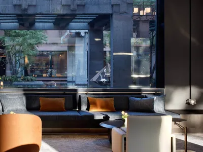 Modern lounge with dark concrete structural beams, large glass walls overlooking a garden, blue-grey sectional sofa with orange accent pillows, and contemporary orange and white lounge chairs.