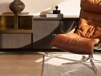 A contemporary living room corner with a brown leather lounge chair, dark wood and brass console, ceramic vase, black sculptural object, and a large framed turquoise ink painting of pine branches on the wall.